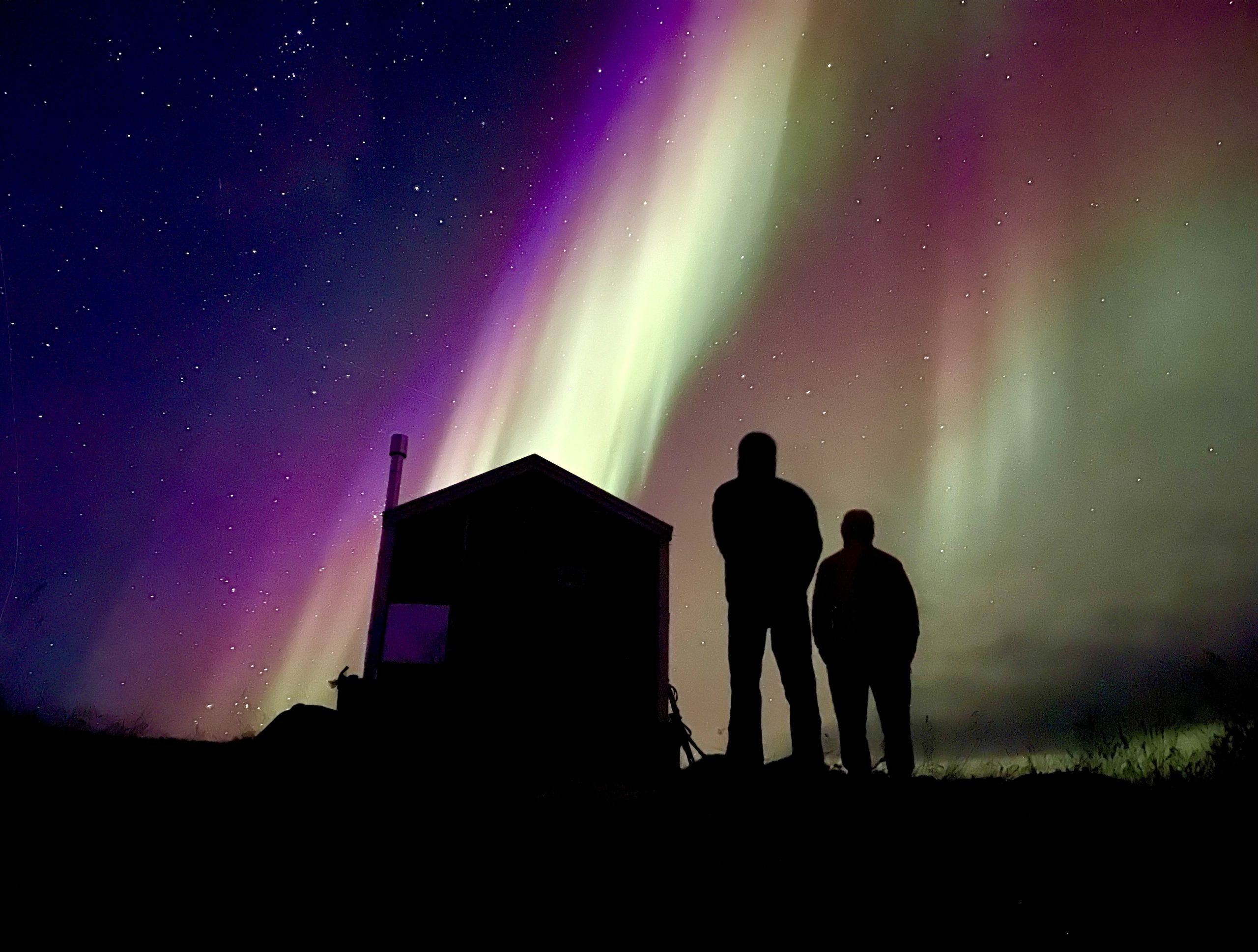 Northern lights over the Arctic Circle Trail in Greenland. Hikers in silhouette standing outside of one of the huts.